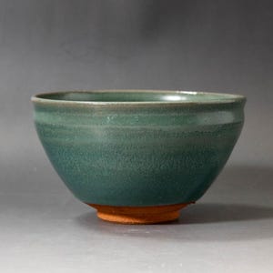May include: A handcrafted teal ceramic bowl with a textured surface and a rounded shape. The bowl has a contrasting orange-brown base. This piece is suitable for serving food or as a decorative item.