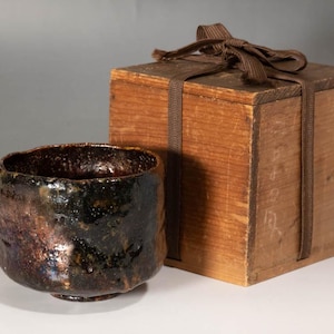 May include: A ceramic bowl with a textured, dark, iridescent glaze. Beside it is a wooden box tied with a brown ribbon. The box has a natural wood finish. The bowl is likely for tea.