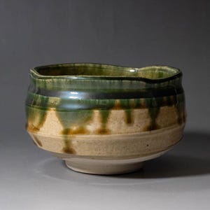 May include: A handcrafted ceramic bowl with an irregular form. The bowl has a two-tone glaze, with a light beige base and a dark green and black rim. The glaze runs down the sides, producing a textured appearance. The bowl rests on a small, white base.