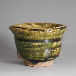 May include: A handcrafted ceramic cup with a unique, textured glaze. The cup features a blend of green, brown, and black hues, with a crackled effect. The design includes horizontal bands of dark color, adding visual interest to this pottery piece.