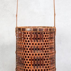 Pode incluir: Cesto cilíndrico de bambu com alça. Apresenta um padrão tecido de tiras de bambu castanho escuro e claro, criando um design geométrico. A alça é feita de bambu curvo. Adequado para armazenamento ou exibição.