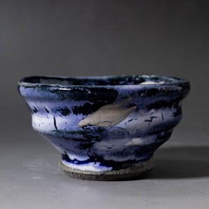 Op de afbeelding: Een keramische kom met een gevlekte glazuur in diep marineblauw, helder wit en lichtpaars. De kom heeft een onregelmatige, golvende rand en een textuurrijke, donkergrijze ongeglazuurde voet. Een enkel, glad, lichtgrijs bladmotief is zichtbaar.