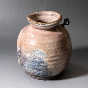 以下が含まれることがあります： 幅の広い厚い縁を持つ丸い陶器の花瓶。ダスティピンク、タン、斑点のあるブルーグレーの色合いの質感のある光沢のある釉薬が施されています。容器の上部側面には、小さな暗い金属製のリングが取り付けられており、表面にはわずかな横方向の隆起が見られます。