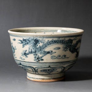 May include: A ceramic bowl with an off-white base color featuring hand-painted blue dragon motifs encircling the exterior. The design includes stylized clouds and wave patterns, with a decorative border near the base and a wide, open rim.