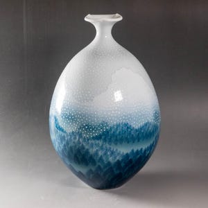 May include: A decorative ceramic vase with a distinctive design. The vase is white at the top, fading into a blue forest-like pattern at the base. The white section has small white dots. The vase has a slender neck and a rounded body.