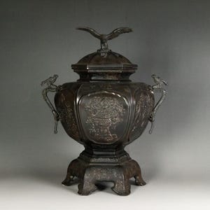 May include: A dark bronze decorative urn with a lid, featuring an eagle finial. The urn has ornate handles with bird details and floral relief patterns. The base is also decorated with floral designs.