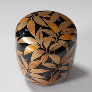 Könnte beinhalten: Eine schwarze, ovale dekorative Box mit Deckel. Die Box ist mit einem goldenen Blattmuster mit Glitzerakzenten verziert. Die Blätter sind in einem sich wiederholenden Muster angeordnet.