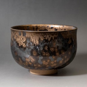 Op de afbeelding: Een keramische kom met een diep, metallic glazuur in een mix van donkerbruine, zwarte en glinsterende bronstinten. Het oppervlak vertoont een gevlekt, kristallijn patroon met druppels en kleurvariaties, rustend op een lichte, effen voet.