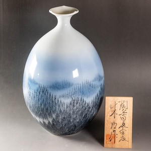 May include: A white and blue ceramic vase with a landscape design. The vase has a gradient of blue and white, with a forest scene painted at the bottom. A wooden plaque with Japanese calligraphy is next to the vase.