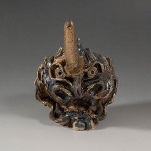 Puede incluir: Una escultura de cerámica de una cara de demonio con cuernos y un cuerno dorado. La cara está detallada con tonos azul oscuro, marrón y beige. La escultura tiene una superficie texturizada y una pequeña abertura en la parte superior del cuerno.