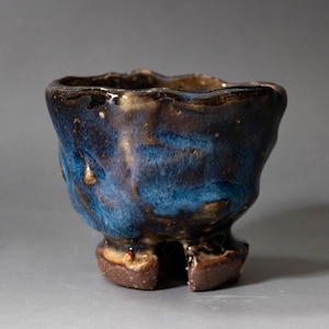 May include: A small, hand-formed ceramic cup featuring a deep blue and dark brown glazed finish. The vessel has an irregular, wavy rim and a textured, uneven surface. It stands on a thick, dark brown base with a distinct vertical cutout notch.