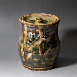 Pode incluir: Um pote cilíndrico de cerâmica com tampa a condizer e um puxador central. A superfície exterior apresenta um vidrado salpicado em tons de verde floresta, castanho terra e creme, com pinceladas verticais abstratas e pontos escuros.