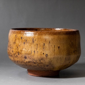 Può includere: Una ciotola in ceramica rotonda con un'ampia apertura e un bordo spesso e curvo. L'esterno presenta una smaltatura color giallo senape terroso con macchie marrone scuro e motivi a goccia verticali, poggiante su una piccola base non smaltata.