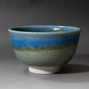 Könnte beinhalten: Keramikschale mit blau-grüner Glasur. Die Schale hat eine breite, abgerundete Form und steht auf einem kleinen, weißen Sockel. Die Glasur hat einen Craquelé-Effekt, der der Oberfläche Textur verleiht. Die Schale wird wahrscheinlich zum Servieren von Speisen oder als Dekorationsartikel verwendet.