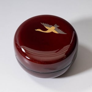 May include: Circular, deep red box with a shiny surface. The lid is decorated with a gold and grey bird in flight. The box is probably made of wood and has a smooth, reflective finish. The design is simple and elegant.