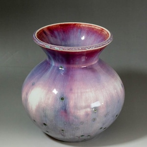 May include: A ceramic vase with a rounded body and a flared neck. The vase has a colour gradient, shifting from deep red at the rim to shades of purple and blue. Small, dark green spots are scattered across the surface.