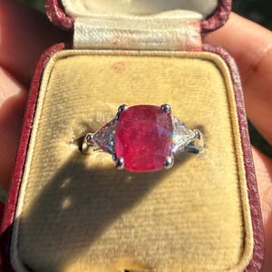 May include: A silver ring featuring a large, cushion-cut ruby, with two smaller, triangular, clear stones either side. The ring is presented in a vintage red jewellery box with a yellow velvet lining.