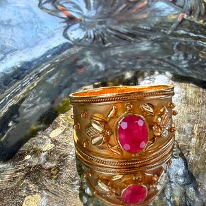 Puede incluir: Un brazalete ancho de tono dorado con una piedra preciosa central de corte ovalado en color rojo rosado vibrante. La superficie metálica presenta motivos florales y de hojas en relieve, enmarcados por bordes finos de estilo cuerda trenzada.