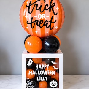May include: A round orange balloon with vertical stripes and the text "trick-or-treat" sits atop a white square box filled with orange and black balloons. The box features white decals of a ghost, bats, a spiderweb, a pumpkin, and "HAPPY HALLOWEEN LILLY".