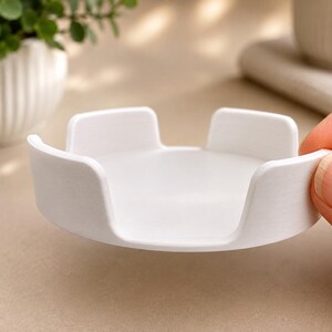 Pu&ograve; includere: Un supporto circolare in plastica bianca con una base piatta e quattro linguette verticali equidistanti che si estendono verso l'alto dal bordo. L'oggetto presenta una finitura opaca liscia e un design semplice e funzionale.