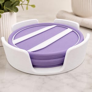Pu&ograve; includere: Una pila di sottobicchieri rotondi viola con strisce diagonali bianche, tenuti all'interno di un supporto circolare bianco. I sottobicchieri presentano una superficie liscia e piatta con un bordo rialzato e il supporto ha quattro sostegni curvi.