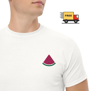Könnte beinhalten: Ein weißes T-Shirt mit kurzen Ärmeln und einer kleinen gestickten Grafik einer Wassermelonenscheibe auf der Brust. Die Wassermelonenscheibe ist rot mit dunkelvioletten Kernen und einer grünen Schale unten, mittig auf der Vorderseite platziert.