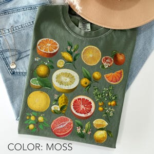 May include: A moss green t-shirt featuring a botanical print of various citrus fruits including oranges, grapefruits, lemons, and limes, displayed as whole fruits, cross-sections, and small branches with leaves and white blossoms.