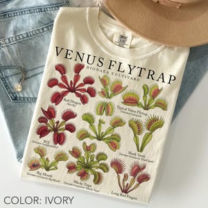 May include: An ivory t-shirt featuring a botanical illustration of various Venus flytrap cultivars. The text reads VENUS FLYTRAP DIONAEA CULTIVARS, with labels for Red Dragon, Typical, Shark Teeth, Big Mouth, Wacky Traps, and Long Red Fingers.