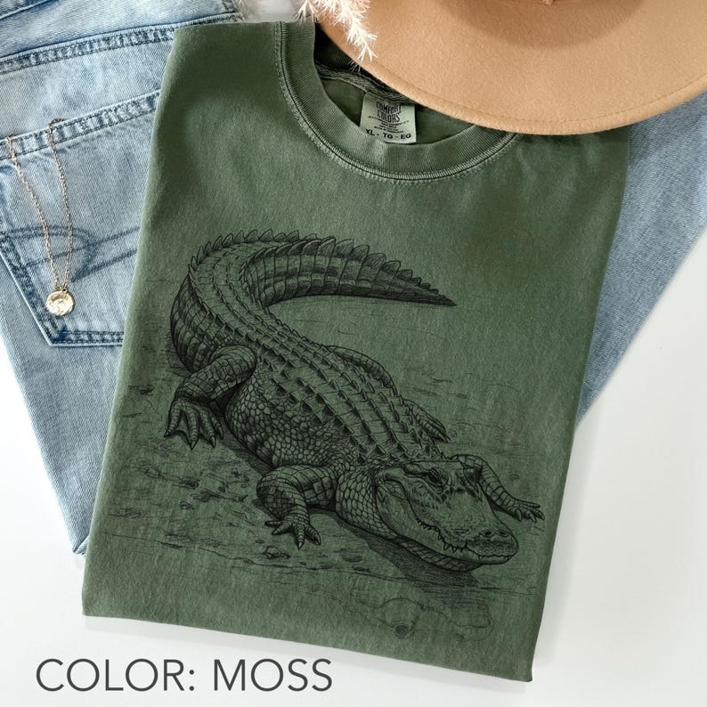 May include: Moss green t-shirt featuring a detailed illustration of an alligator. The shirt is a crew neck style and is paired with blue jeans and a necklace. The text "COLOR: MOSS" is at the bottom of the image.