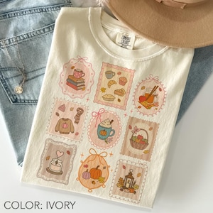 Puede incluir: Camiseta de color marfil con una serie de ilustraciones con temática otoñal en marcos decorativos. El diseño incluye calabazas, café, productos horneados y hojas. La camiseta se muestra con vaqueros azules y un collar. El texto dice "COLOR: IVORY".
