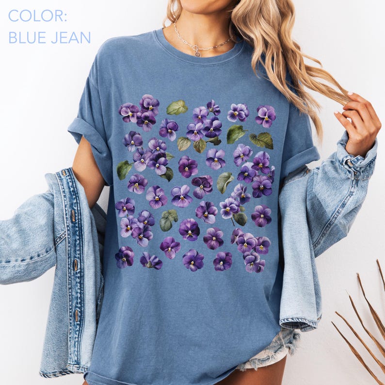 May include: Blue jean colored t-shirt featuring a watercolor floral design of purple pansies and green leaves. The text "COLOR: BLUE JEAN" is in the upper left corner. The shirt has rolled-up sleeves.
