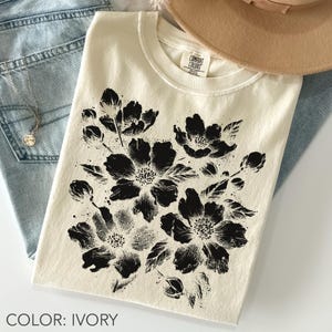 以下が含まれることがあります： 大きな黒い花柄が特徴のアイボリー色のTシャツ。クルーネックのシャツは柔らかい素材で作られています。「COLOR: IVORY」の文字が画像の下部にあります。シャツはブルージーンズとネックレスと組み合わせています。