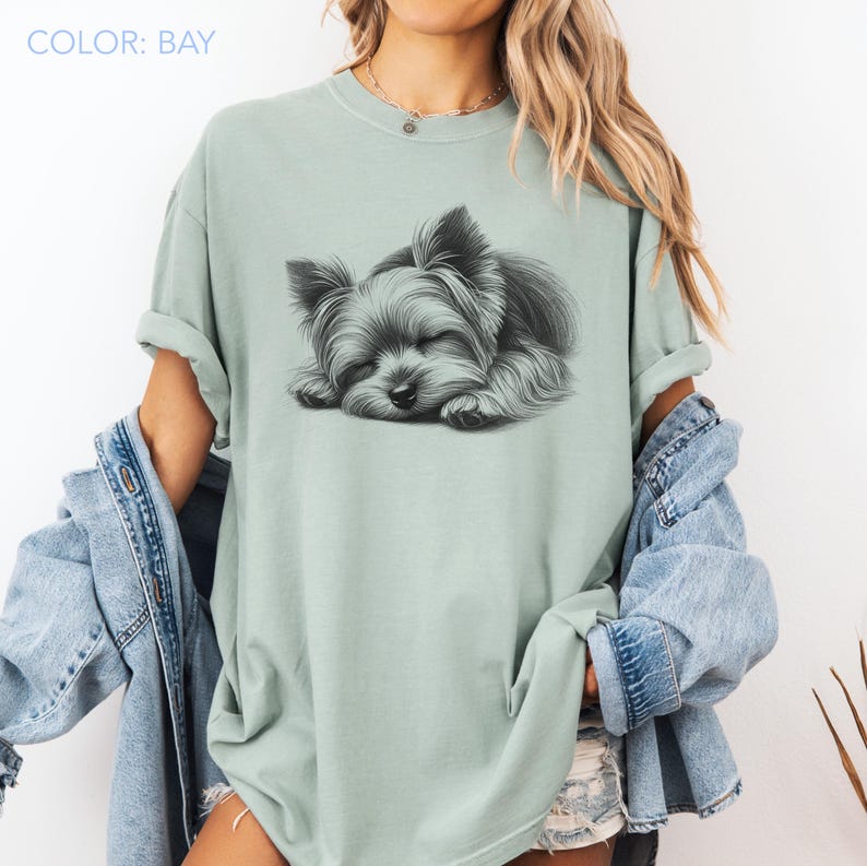 Puede incluir: Camiseta verde claro con un gr&aacute;fico de un Yorkshire Terrier durmiendo. El texto "COLOR: BAY" est&aacute; en la esquina superior izquierda. La camiseta tiene mangas enrolladas y un ajuste relajado.