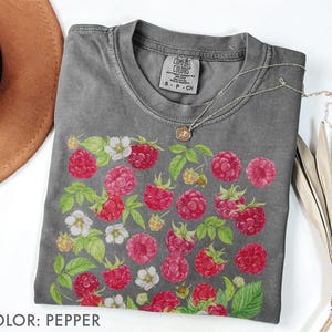 Op de afbeelding: Pepperkleurig T-shirt met een ronde hals. Het shirt heeft een aquarelontwerp van rode frambozen, witte bloemen en groene bladeren. Een zilveren ketting met een hanger ligt op het shirt. Het label op het shirt luidt "Comfort Colors".