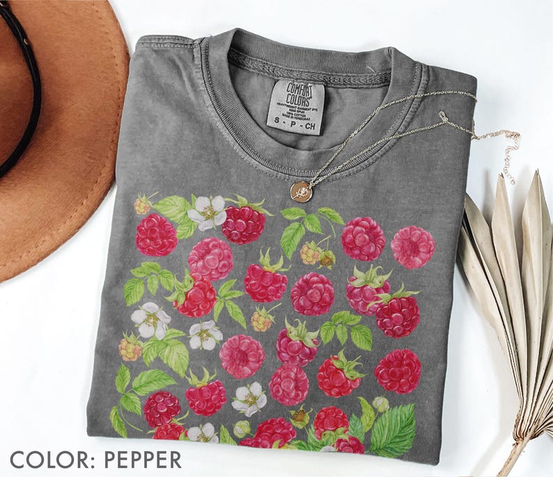 Op de afbeelding: Pepperkleurig T-shirt met een ronde hals. Het shirt heeft een aquarelontwerp van rode frambozen, witte bloemen en groene bladeren. Een zilveren ketting met een hanger ligt op het shirt. Het label op het shirt luidt "Comfort Colors".
