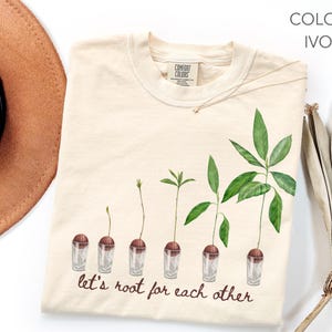 May include: Ivory-coloured t-shirt with a watercolour illustration of plant growth stages in small glasses. The text "let's root for each other" is written in a script font. The shirt is a Comfort Colors brand.