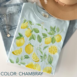 May include: A light blue t-shirt with a lemon and leaf design. The shirt features a watercolor illustration of yellow lemons, green leaves, and white flowers. The text "COMFORT COLORS" is on a tag. The color of the shirt is chambray.