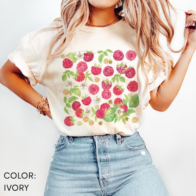 Op de afbeelding: Ivoor kleurig T-shirt met een aquarel ontwerp van rode frambozen, groene bladeren en witte bloemen. Het ontwerp is gecentreerd op de voorkant. De tekst "COLOR: IVORY" staat linksonder in de afbeelding.