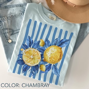 May include: Light blue t-shirt with a lemon design. The shirt features a watercolor illustration of a sliced lemon, a whole lemon, and blue leaves. Vertical blue stripes are on the shirt. The text "COLOR: CHAMBRAY" is at the bottom of the image.