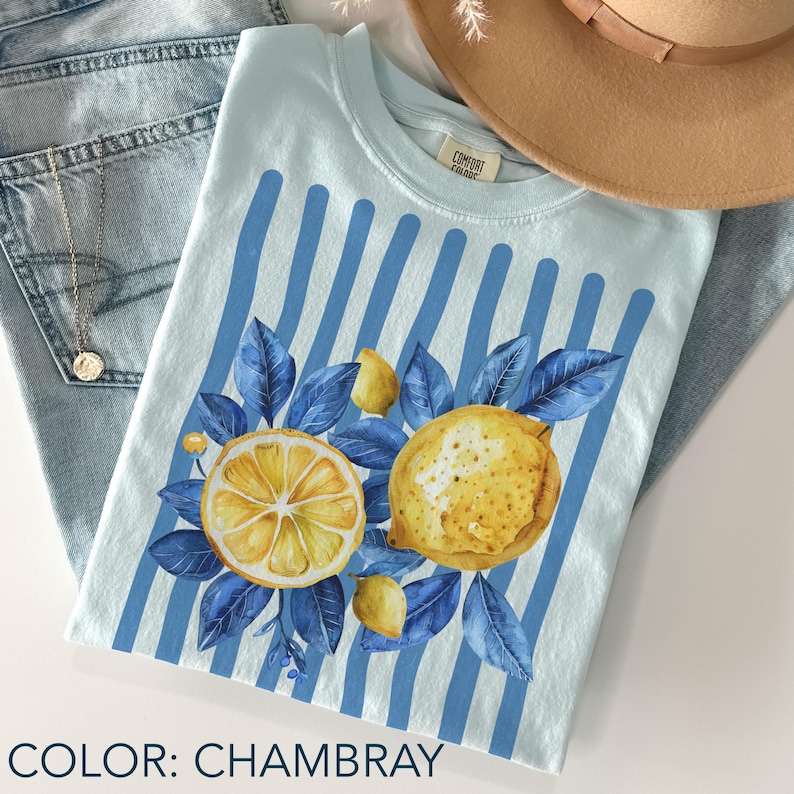 May include: Light blue t-shirt with a lemon design. The shirt features a watercolor illustration of a sliced lemon, a whole lemon, and blue leaves. Vertical blue stripes are on the shirt. The text "COLOR: CHAMBRAY" is at the bottom of the image.
