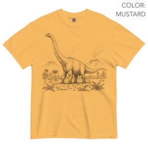 Pu&ograve; includere: T-shirt color senape con un'illustrazione dettagliata di un dinosauro Brachiosaurus in un paesaggio preistorico. Il design include palme, vulcani e una rappresentazione dettagliata del dinosauro.