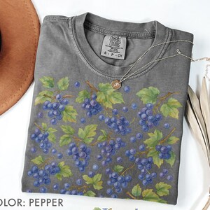 May include: A gray Comfort Colors t-shirt featuring a watercolor design of purple grapes and green leaves. The shirt has a crew neck and short sleeves. A silver necklace with a pendant is draped on the shirt. The color of the shirt is pepper.
