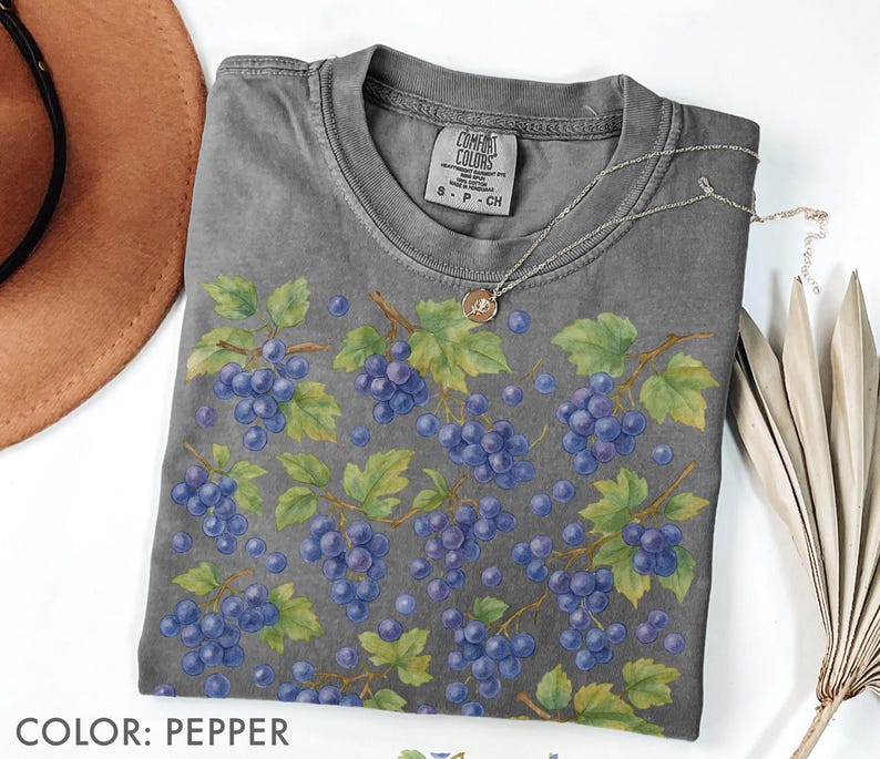 May include: A gray Comfort Colors t-shirt featuring a watercolor design of purple grapes and green leaves. The shirt has a crew neck and short sleeves. A silver necklace with a pendant is draped on the shirt. The color of the shirt is pepper.