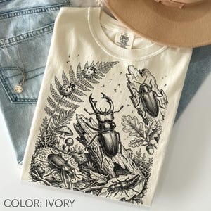 Può includere: T-shirt color avorio con un'illustrazione dettagliata in bianco e nero di vari insetti, tra cui un cervo volante, coccinelle e altri coleotteri, ambientati in una scena naturalistica con felci e funghi. La maglietta ha un girocollo e il testo "COLOR: IVORY" in basso.