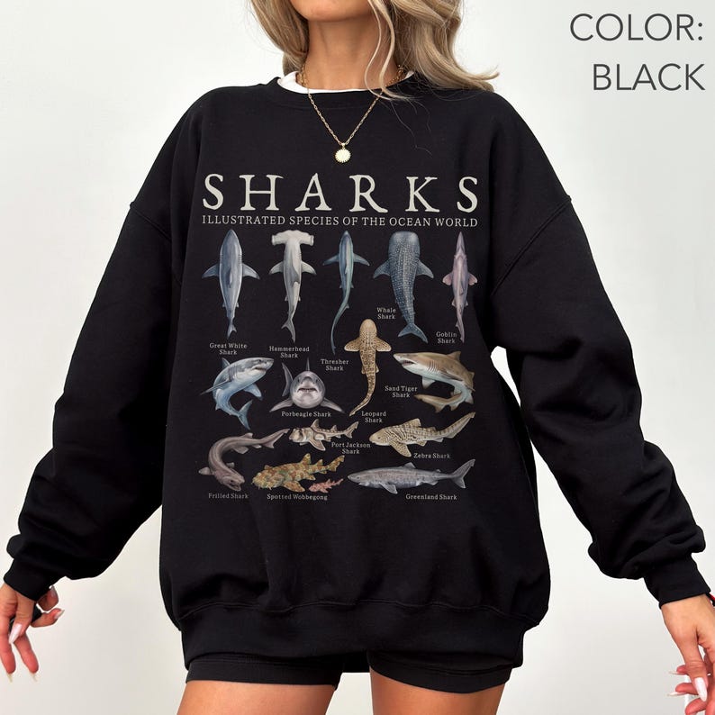 K&ouml;nnte beinhalten: Ein schwarzes Langarm-Sweatshirt mit einem Grafikdruck mit der Aufschrift SHARKS ILLUSTRATED SPECIES OF THE OCEAN WORLD. Das Design zeigt verschiedene Haiarten mit ihren Namen unter jedem Tier, darunter der Wei&szlig;e Hai und Hammerhai.