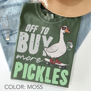 Könnte beinhalten: Moosgrünes T-Shirt mit dem Text "OFF TO BUY more PICKLES" und einer Grafik einer Gans mit Sonnenbrille und roten Stiefeln auf einem Skateboard, die ein Glas Gurken hält. Das T-Shirt ist auf einer Jeans gefaltet.
