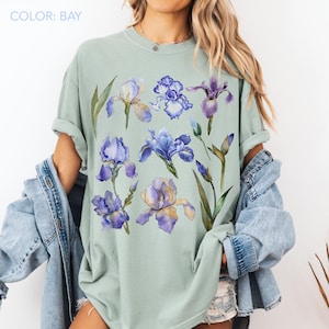 Könnte beinhalten: Ein beerenfarbenes Kurzarm-T-Shirt mit einem Grafikdruck aus verschiedenen violetten und blauen Irisblüten mit grünen Stielen und Blättern. Das T-Shirt hat einen Rundhalsausschnitt und eine lockere Passform, getragen unter einer blauen Jeansjacke.