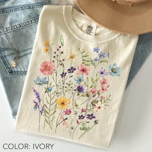 Puede incluir: Camiseta de color marfil con un dise&ntilde;o floral en acuarela con flores rosas, azules, amarillas y moradas. La camiseta est&aacute; doblada, con un sombrero y unos vaqueros en el fondo. El texto "COLOR: IVORY" est&aacute; en la parte inferior.