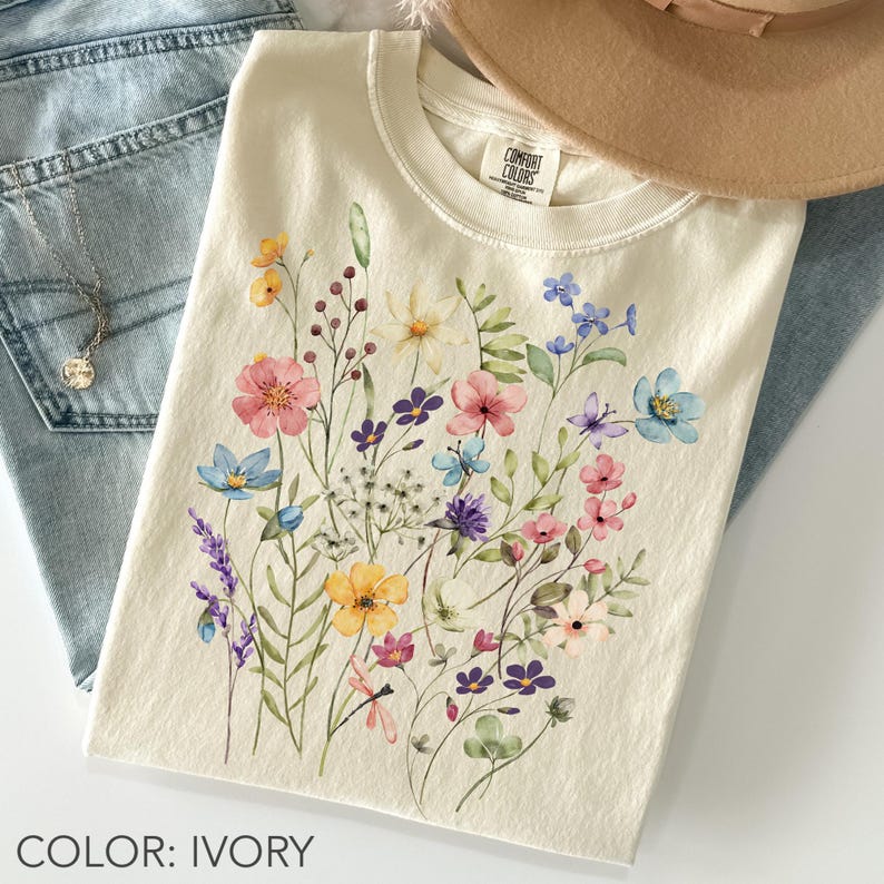 Puede incluir: Camiseta de color marfil con un dise&ntilde;o floral en acuarela con flores rosas, azules, amarillas y moradas. La camiseta est&aacute; doblada, con un sombrero y unos vaqueros en el fondo. El texto "COLOR: IVORY" est&aacute; en la parte inferior.