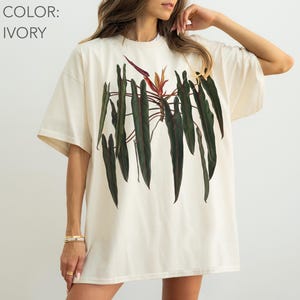May include: An ivory short-sleeve t-shirt featuring a large, centered botanical illustration of a philodendron plant with long, dark green, pointed leaves and a single reddish-orange flower bud at the top center of the plant graphic.